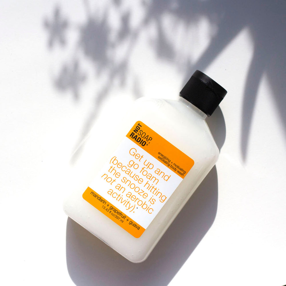 A mandarin orange colored body scrub flat lay on a white background with leaf shadows in the background.