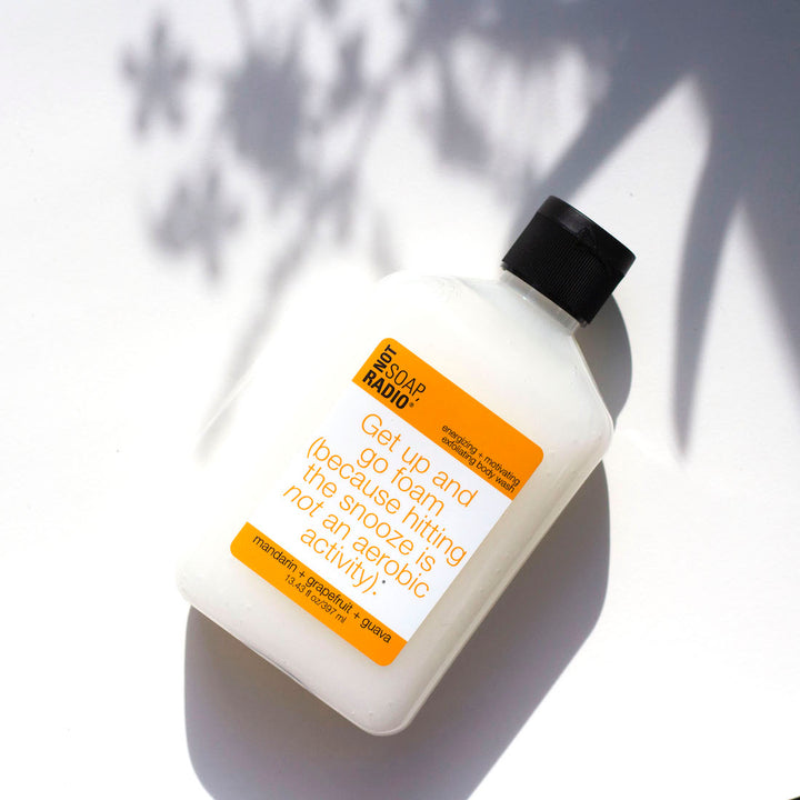 A mandarin orange colored body scrub flat lay on a white background with leaf shadows in the background.