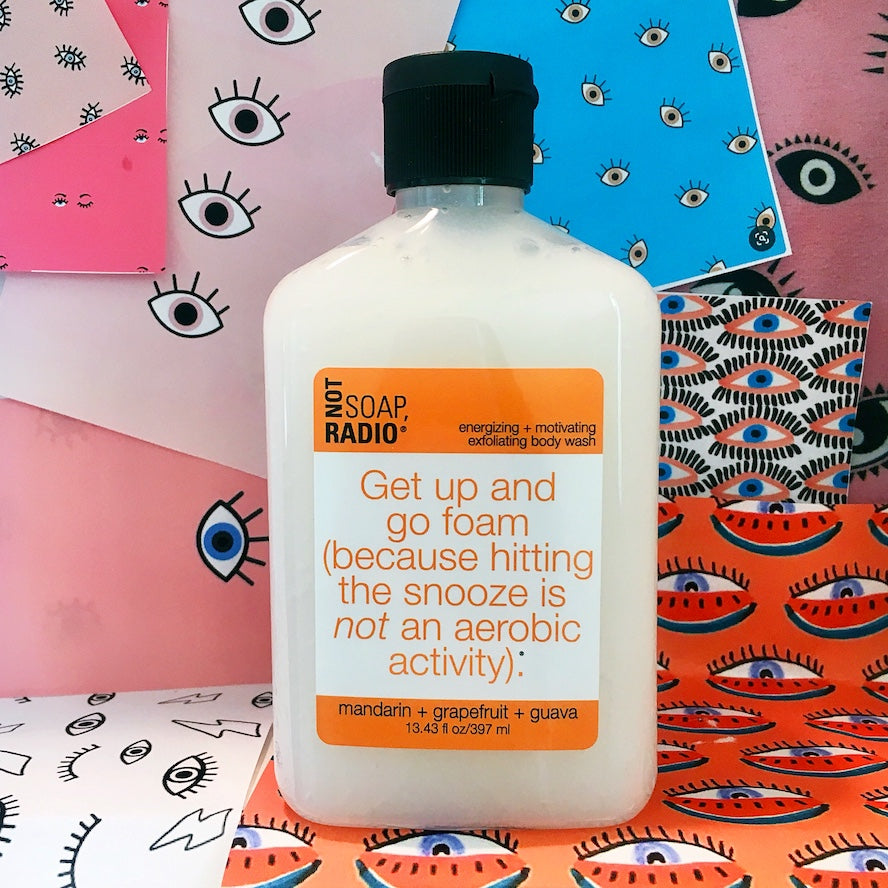 An orange labeled body scrub in front of a collage with eye themed cutouts.