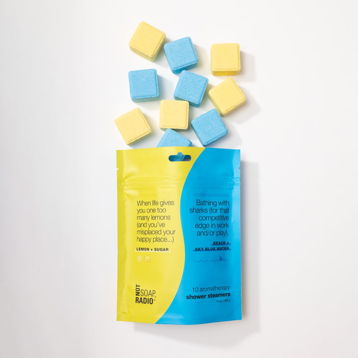 Flatlay of Bathing with sharks + When life gives you one too many lemons variety pack Shower Steamers pouch, with blue and yellow shower steamer tablets coming out of bag