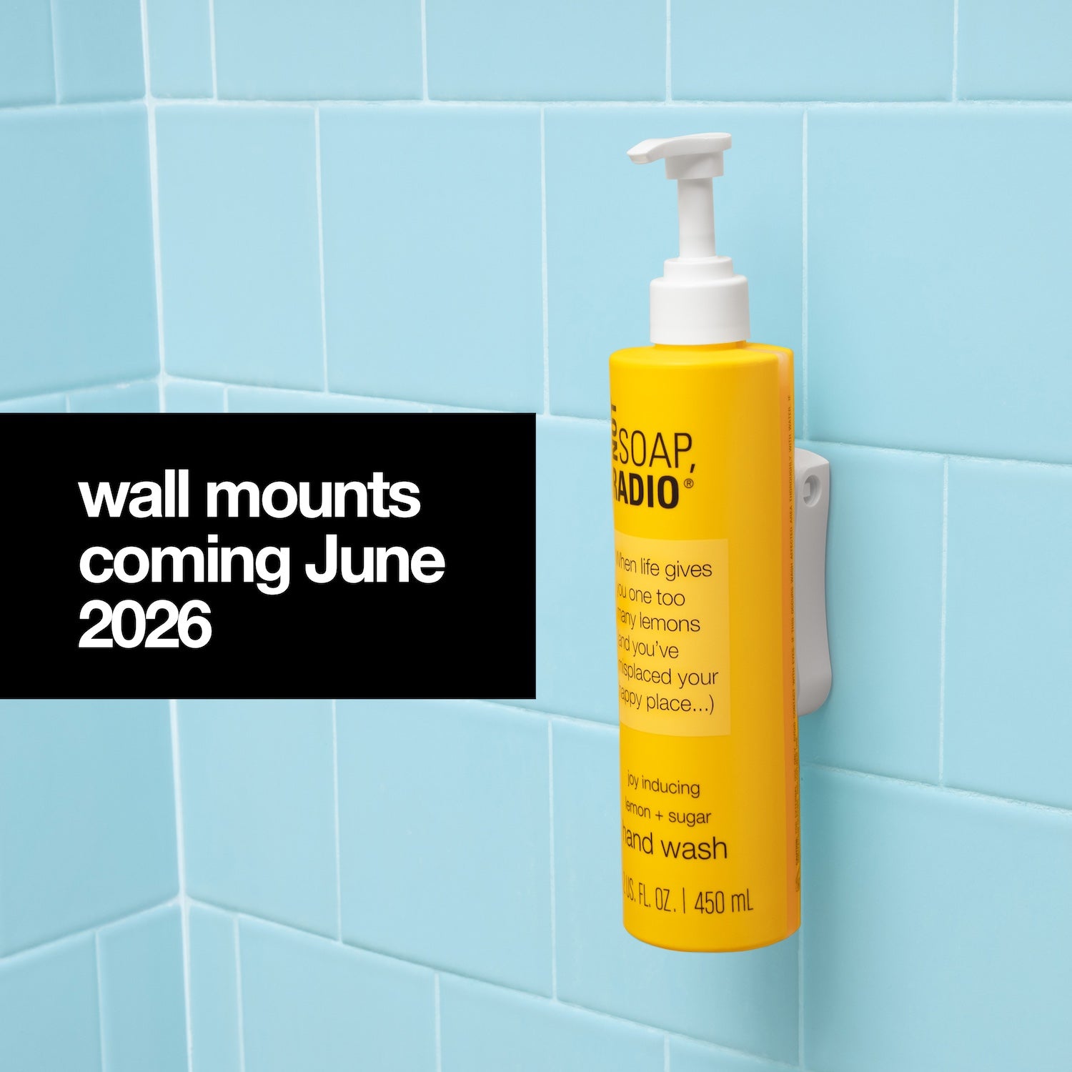 A blue tile shower background with a black box on the left hand side containing white text stating "wall mounts coming June 2026". To the right of the black text box a yellow Not Soap Radio hand wash bottle with a white pump cap on top and a white adhesive wall mount on the back of the bottle mounting it to the wall.