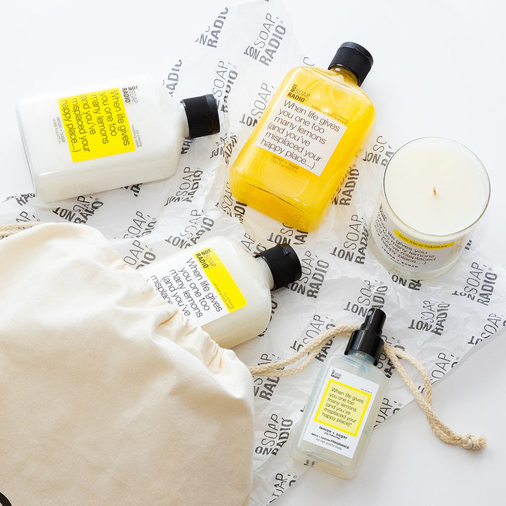 A lemon and sugar scented body lotion, body mist, shower gel, candle, and exfoliating scrub coming out of a drawstring bag.