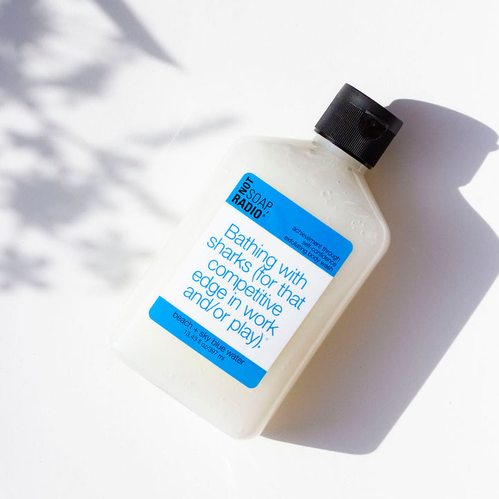 A blue themed exfoliating body wash flat lay on a white background with leaf shadows.