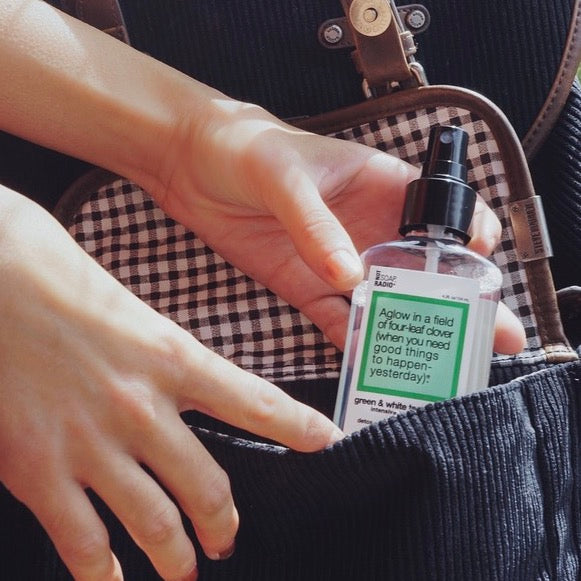 A green clover scented body mist being placed inside of a satchel bag.