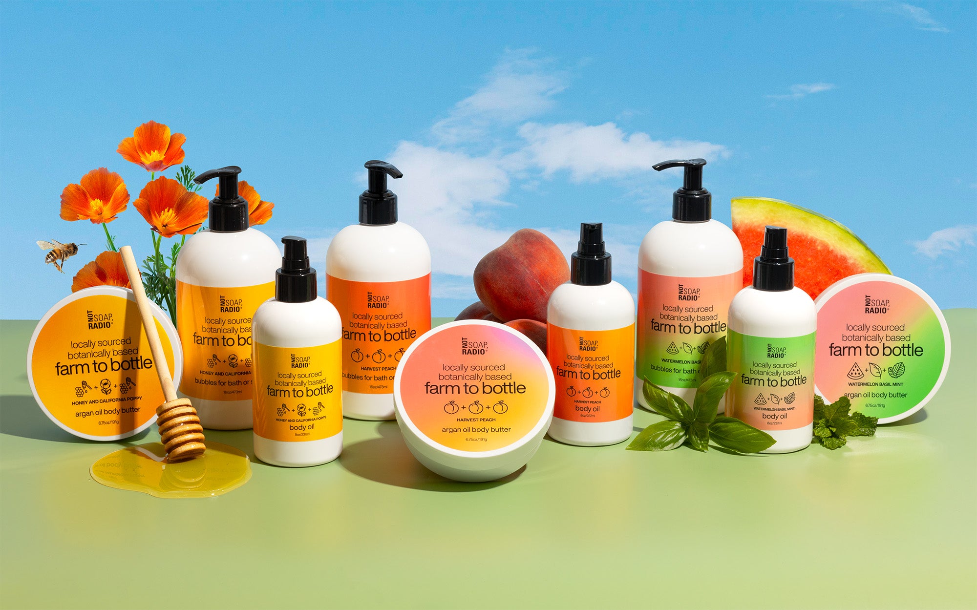 The farm to bottle bath and body collection - all bottles in a row with ingredients behind them, includinga honey bee and california poppy flower, harvest peaches and watermelon basil and mint.