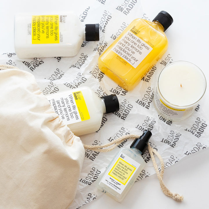 Lemon and sugar scented lotion, exfoliating scrub, candle, body mist coming out of a Not Soap Radio drawstring bag.