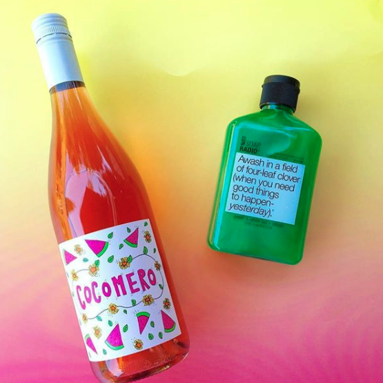 A green bubble bath gel laying next to a watermelon wine on top of a pink to yellow ombre background.