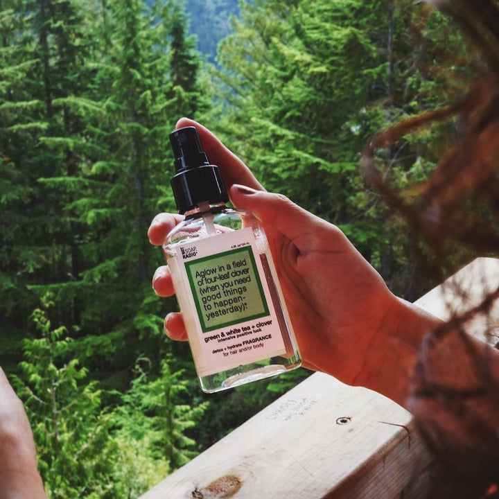 A green clover scented body mist being held up in front of a green forest.