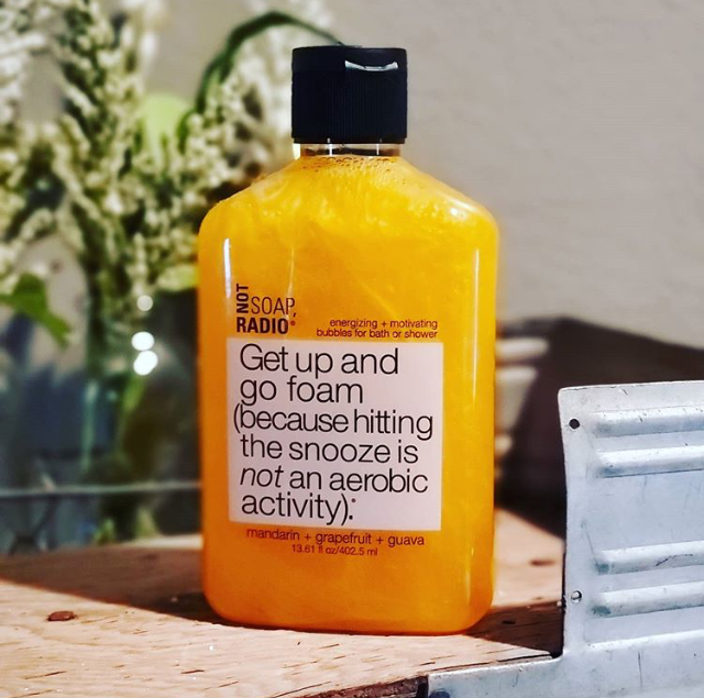 An orange bath and shower gel standing on top of a wooden plank with plants in the background.