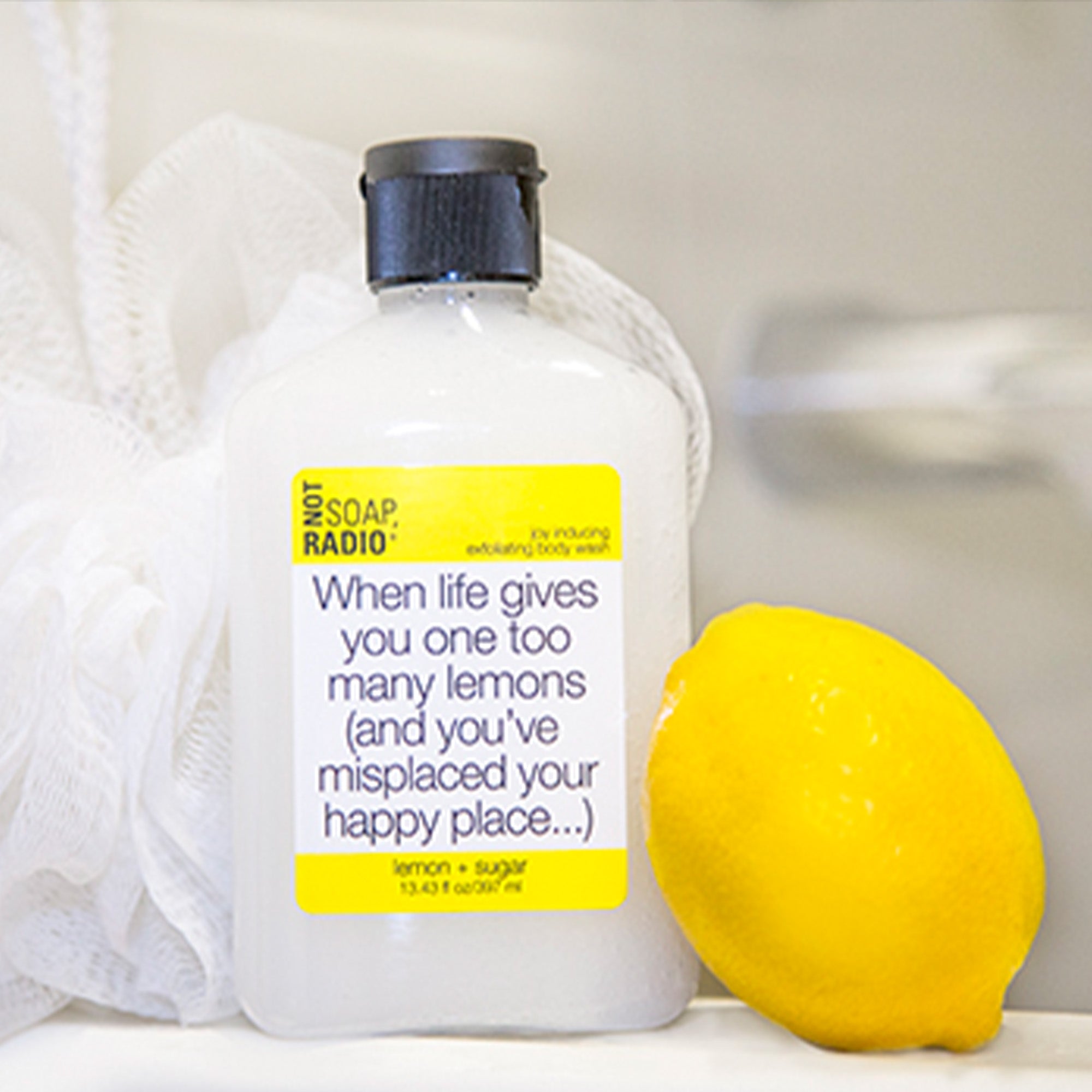 lemon sugar scented exfoliating body wash, on the edge of a tub with a lemon and loofah.
