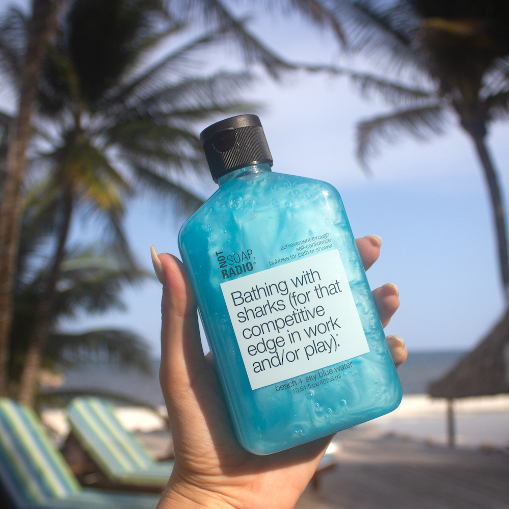 A beach and blue water scented bubble bath gel being held in front of palm trees on a beach.