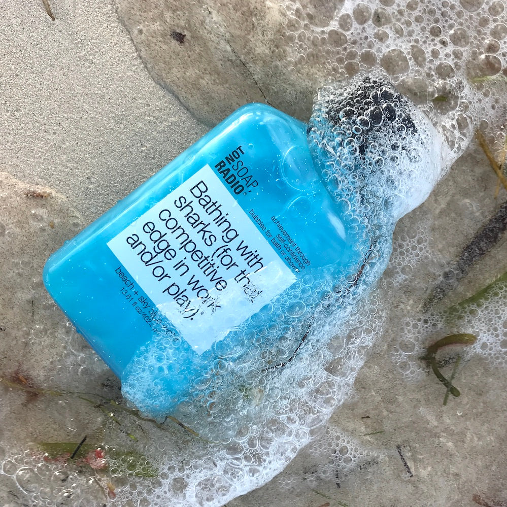 A beach and sky blue water scented bubble bath gel laying on top of sand with an ocean wave going over the bottle.
