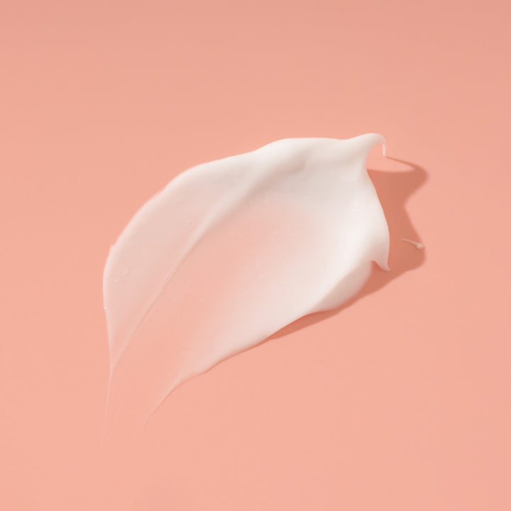 Product swatch of white body butter formula, rich in Argan oil, shea butter, and avocado oils - photographed on a peach pink background