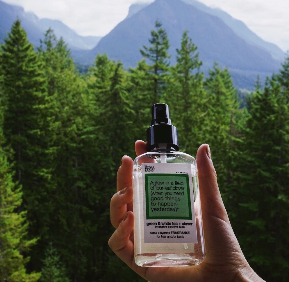 A green clover scented body mist being held in front of a view of green trees and mountains.