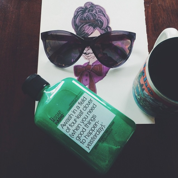 A lifestyle flatlay of a bubble bath gel next to a cup of coffee and sunglasses.