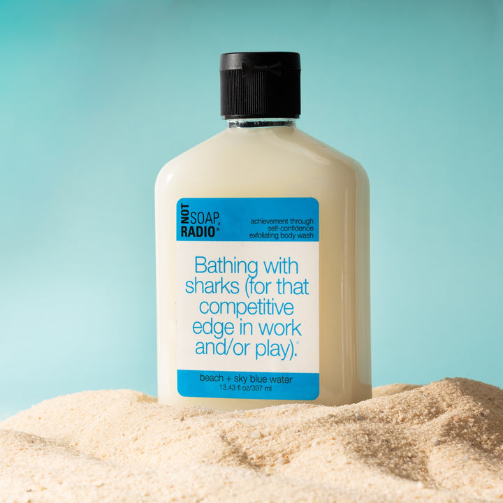 Beach scented exfoliating body wash standing in a pile of sand on a blue background