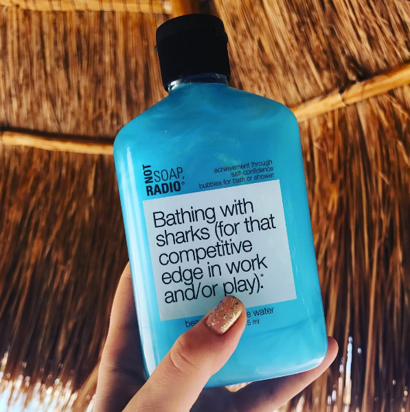 A light blue bath and shower gel being held in front of a palm leaf gazebo.