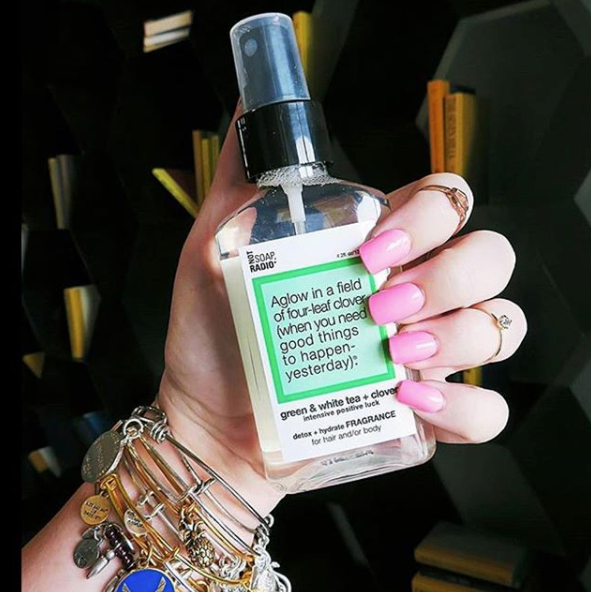 A woman's hand with pink nails holding a green tea and clover scented hair and body fragrance mist.